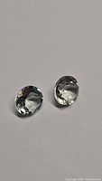 Two oval natural prasiolite stones, diamond cut, shown side by side on gray background with clear light green color and high clarity.
