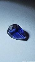 Close-up photo of a deep purple pear-shaped faceted iolite gemstone on a white surface with good lighting highlighting its color and cut.