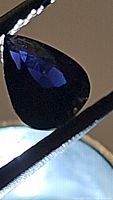 Close-up photo of the teardrop-shaped deep dark purple Spinel gemstone held by tweezers under bright light, showing facets and clarity.