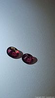 Pair of oval cut natural red spinel gemstones displayed on a white background under diffuse lighting showing color and clarity.