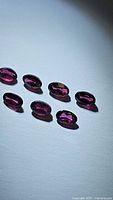 Seven oval cut rhodolite garnet gemstones arranged on a white surface, showing their size, shape and color.