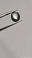 Close-up photo of natural oval cut green blue sapphire held with tweezers, showing the gemstone's color and faceted surface.
