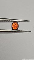 Oval cut orange Spessartite garnet held by tweezers showing clarity and cut facets.