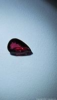 Close-up image of a pear-shaped deep red ruby gemstone on a light background, showing facet reflections and shape clearly.