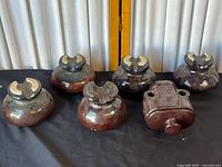 Six ceramic electrical insulators on black surface with dark and reddish-brown two-tone glaze. Five insulators are rounded with sculpted crescent tops; one insulator is rectangular with embossed text and two large holes.