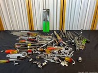 Wide shot showing an assortment of hand tools including wrenches, screwdrivers, sockets, sockets, drill bits, saws, hex keys scattered on a black cloth.
