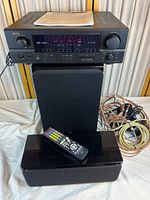 Front view of Denon AVR-1705 AV receiver on top of NHT speaker with another speaker in front, remote control on speaker, speaker wires coiled on right, and manual on receiver.