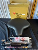 Photo showing the black motorcycle-style seat, metal base/legs, mounting hardware, and Gibraltar stickers arranged on black cloth in front of the original box.