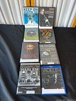 Nine music DVDs and Blu-ray discs shown posed on black cloth, visible titles include Guns N' Roses, Adele, The Chieftains, Benny Goodman, Grateful Dead, Keep On Keepin' On, Dear Jerry, A Great Day in Harlem