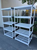 Two white plastic shelving units with five shelves each and black corner reinforcements, shown outdoors on pavement with greenery in background.