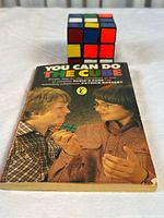 Front cover of 'You Can Do the Cube' book alongside the Rubik's Cube puzzle placed behind it, showing both items clearly.