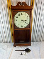 Full view of antique Seth Thomas wooden wall clock showing clock face, wood case with decorative carved top, glass front panel, pendulum and winding keys in front.