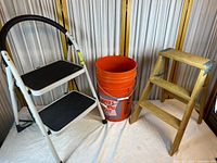 Image showing wood and metal step ladders next to a Homer’s orange 5-gallon bucket on white and wood background.