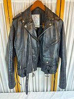 Front view of black leather jacket on wooden hanger showing wide collar, zippered sleeves, epaulettes, front zipper and snap flap pocket with belt at bottom.