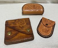 Photo showing all three leather cases on a white background: a wallet, a small pouch, and an eyeglass/pen case with floral tooled designs.