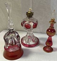 Three vintage glass items displayed on white fabric: a cranberry glass bell, an oil lamp with brass burner, and a decorated perfume bottle.