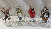 Four clear vintage glasses featuring colorful Popeye characters Olive Oyl, Rough House, Brutus, and Wimpy, standing in a row showing front character graphics.