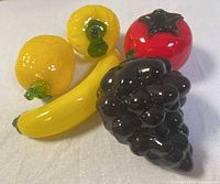 Five colorful vintage hand-blown glass fruit figurines grouped on white cloth, including banana, two lemons, grape cluster, and tomato.