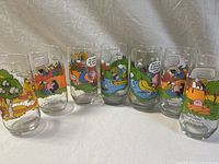 Full set of 7 clear tall glasses arranged in a row, each decorated with colorful Peanuts Camp Snoopy camping scenes.