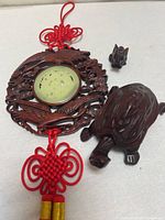 Photo showing carved wooden hanging medallion with green inset disc and red decorative knots, alongside the dark wooden turtle and small elephant figures on a flat surface.