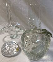 Photo showing the clear glass basket with handle at back, clear apple with bubbles and green leaf in front left, snowflake paperweight front left.