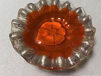 Top and angled view of hand-blown art glass bowl showing vibrant orange interior and clear crackle-effect ruffled rim with thick glass base.
