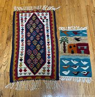 Lot image showing two handwoven wool rugs on wooden floor. Left rug has tribal geometric pattern with fringes; right rug has folk art design with birds, houses, and palm tree with fringes.