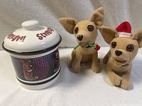Photo showing three items: two plush Taco Bell chihuahuas with one wearing a red Santa hat and the other with a green holiday collar, placed beside a ceramic Scooby-Doo themed condiment jar with lid