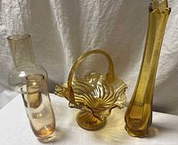 Four pieces include a ribbed amber glass basket with ruffled edge and handle, a tall swung-style amber vase with petal top, a modernist small amber bud vase, and a clear glass bulbous vase with narrow neck and flared rim.