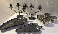 Photo showing two ornate silver-tone 3-arm candelabras, two leaf-shaped pewter trays, and five stainless steel tumblers with medallion accents.