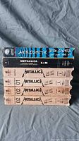 Photo showing all six VHS tapes stacked with titles visible: Aerosmith box on top, multiple Metallica tapes below with varying spine designs showing the live show locations and numbering.