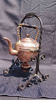 Front right side view of the copper kettle on wrought iron stand showing the ornate design, twisted brass handles, and burner.