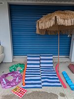 Full set of beach day items including palm tree umbrella, two foldable beach lounge seats, pool noodles and floaties, and a rolled plastic tablecloth.