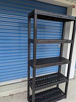 Full view of black plastic 5-tier shelving unit standing outside in front of blue roller shutter door.