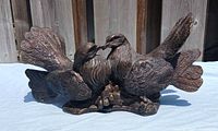 Photo of cast metal (bronze-tone) statue showing two detailed doves touching beaks on naturalistic base, side angle view