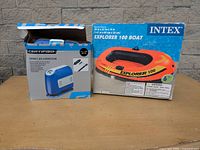 Photo of packaging for Intex Explorer 100 inflatable boat and Certified Compact Air Compressor on table against a brick wall.