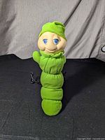 Full front view of the Glo Worm plush nightlight standing upright on a black surface showing the soft green segmented body and cream colored face with blue eyes and a green hat.