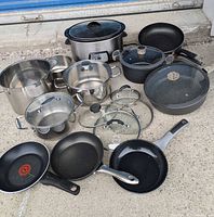 Wide view of pots, pans, lids and slow cooker displayed outdoors on concrete.