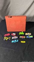 Orange plastic fold and carry Matchbox case with 11 assorted toy cars in front of it on a black cloth surface.