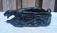 Side view of glossy black ceramic black panther planter resting on white cloth with wooden fence background.