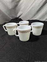 Four Pyrex milk glass mugs displayed on a dark cloth background, showing brown floral border pattern near the rim.