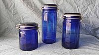 Three cobalt blue glass jars in small, medium, and large sizes with metal clamp lids and rubber seals, shown standing upright on a fabric surface.