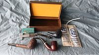 Three vintage smoking pipes, pipe cleaning accessories, metal pipe tool, and wooden box with yellow interior lining