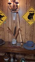 Display on wood panel wall showing two Akubra hats with feathers on table, 3 wooden boomerangs in a ceramic container, a didgeridoo standing behind, and yellow metal signs on wall.