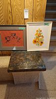 Photo showing two framed art pieces placed on a stone table against wooden paneling and stairs: one is a vintage yellow flower embroidery (16x13), the other is a framed painting (9x12) with abstract figures in reddish background and a signature.
