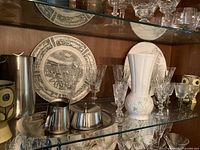 Shelf display showing Wedgwood and Royal Worcester collector plates, crystal wine and aperitif glasses, metal creamer and sugar bowl with tray, and Belleek vase.