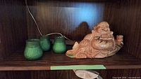 Photo of a ceramic Laughing Buddha statue in brownish glaze sitting on a wooden shelf next to three green glass candle holders with embossed patterns.