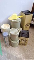 Overall shot showing group of vintage metal and plastic kitchen canisters including two large cream Ekco canisters with marbled finish, beige Tupperware canisters, tall beige metal canisters with floral artwork, and yellow plastic scoop.