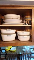 Wooden cabinet shelf with stacked white Corning Ware casserole dishes and bowls, metal stands to the right