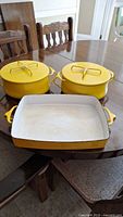 Two yellow Dansk enamelled pots with lids and one large yellow rectangular enamelled baking pan on dining table.